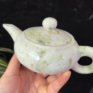 Puede incluir: Una pequeña tetera de color jade con un cuerpo redondeado, un pico y un asa. La tapa tiene un pequeño pomo redondo. La tetera es de color verde pálido con vetas verdes más oscuras. La tetera se sostiene en una mano.