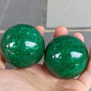 Könnte beinhalten: Zwei grüne Jadekugeln, auch bekannt als Baoding-Kugeln, werden in einer Hand gehalten. Die Kugeln sind glatt und poliert, mit einem dezenten Marmormuster.
