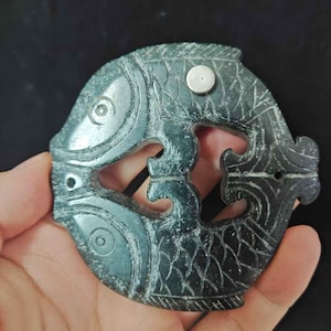 May include: Dark green jade pendant carved in the shape of two fish intertwined. The fish have detailed scales and eye features. A small, silver-colored metal piece is attached to the top. The pendant is held in a hand.