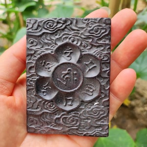 May include: A dark brown, rectangular pendant with intricate carvings. The design features a central floral motif surrounded by stylized symbols and a swirling cloud-like pattern. The pendant is held in a hand, showcasing its detailed texture.