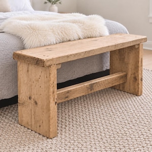May include: A rectangular wooden bench constructed from light brown, weathered timber with a visible grain and natural knots. The bench features a flat seat supported by two thick, solid vertical legs connected by a horizontal stretcher bar.