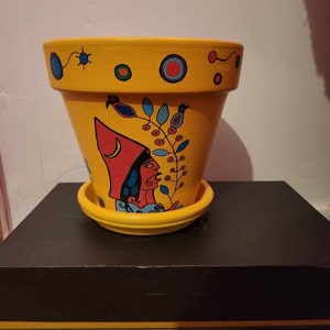 Könnte beinhalten: Ein gelber Terrakotta-Blumentopf mit handbemaltem Design, das eine Frau in einem roten Hut, blaue und grüne Blätter sowie rote und blaue Kreise zeigt. Der Topf hat eine passende Untertasse.