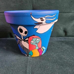 Peut inclure: Un pot en céramique bleu avec un motif peint à la main représentant des personnages du film L'Étrange Noël de monsieur Jack. Les personnages incluent Jack Skellington, Sally et Zero le chien fantôme.