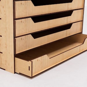 May include: A wooden organizer with five drawers, one of which is open. The drawers are made of light wood with dark brown trim. The organizer is sitting on a white surface.