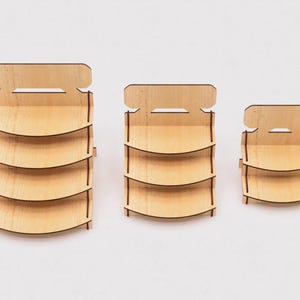 May include: Three wooden display stands with three shelves each. The stands are different sizes, with the largest stand on the left and the smallest stand on the right. The stands are made of light brown wood and have a simple design.