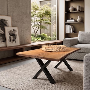 May include: A square wooden coffee table with a natural wood finish and black metal X-shaped legs. A woven basket sits on top. The table is placed on a beige rug in a modern living room setting.