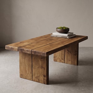 May include: A rectangular wooden coffee table with a natural finish. The table has a planked top and sturdy, block-style legs. A dark bowl filled with moss and two books rest on the table's surface.