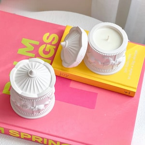 Op de afbeelding: Twee witte keramische kaarshouders in de vorm van een draaimolen met een witte kaars in een van de houders. De kaarshouders staan op een roze en geel boek met het woord "vreugde" op de kaft.