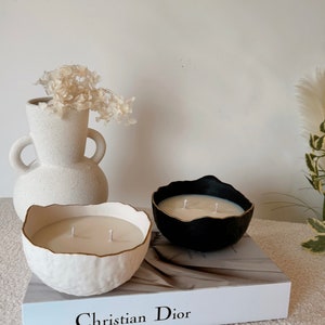 Puede incluir: Dos velas decorativas en recipientes de cerámica blanca y negra con bordes dorados. Las velas están sobre un libro blanco con el texto "Christian Dior" impreso en la portada.