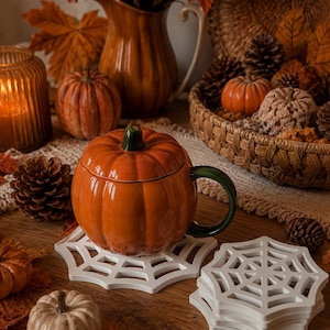 Può includere: Una tazza in ceramica arancione lucido a forma di zucca con coperchio e manico verde scuro, appoggiata su un sottobicchiere bianco a forma di ragnatela. Una pila di sottobicchieri identici si trova accanto, circondata da piccole zucche e pigne.
