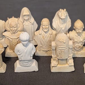 Könnte beinhalten: Eine Sammlung von 12 Star Wars Charakterbüsten, in Weiß- und Brauntönen bemalt. Die Büsten werden auf einzelnen Sockeln präsentiert und zeigen Charaktere wie Darth Vader, Yoda und R2-D2.