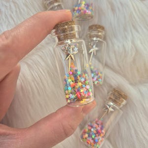 May include: Small glass bottles with cork stoppers, filled with colourful star-shaped confetti. Each bottle has a silver star charm inside. The bottles are displayed on a fluffy white surface.