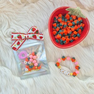 Peut inclure: Une collection de perles et de bijoux sur le thème de la fraise. Un bol rouge en forme de fraise est rempli de perles orange et vertes. Un bracelet avec le nom "ERIN" est à côté d'un sachet de perles avec un nœud fraise.