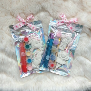 Puede incluir: Dos bolsas transparentes iridiscentes con cremallera atadas con cintas rosas, cada una con un kit de manualidades para pintar. Los kits incluyen botes de pintura, purpurina, pinceles y figuras de cerámica de un unicornio y un ángel.
