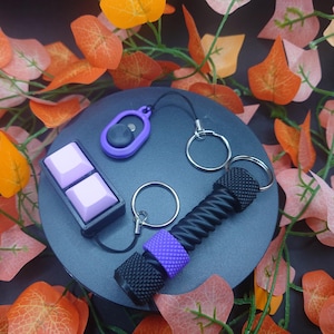 May include: A set of purple and black keychains with a purple and white button, a black and purple twisty keychain, and a black and silver key ring.