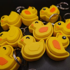 May include: A group of yellow rubber duck keyrings with orange beaks and feet. The keyrings are in the shape of a duck and have a ring attached to the top.