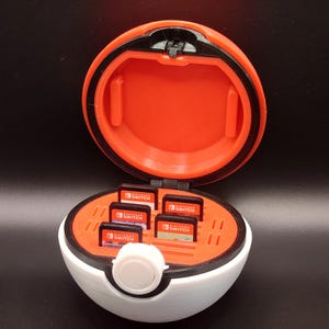 May include: A white and red Poke Ball shaped case with a black lid that opens to reveal slots for Nintendo Switch game cartridges. The case has a white button on the front.