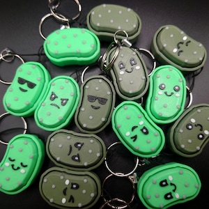May include: A selection of novelty keyrings with pickle designs. The keyrings are in shades of green and olive, with various facial expressions. Each keyring has a silver ring attached.