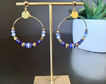 May include: A pair of gold hoop earrings with blue and white beads. The earrings are hanging from a gold stand.