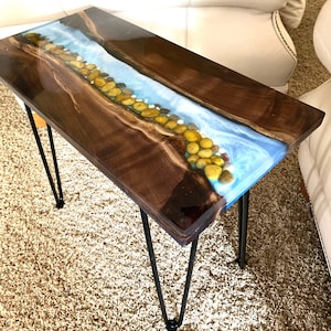May include: A wooden table with a blue and white epoxy river design and embedded stones. The table has black hairpin legs and is sitting on a beige rug.
