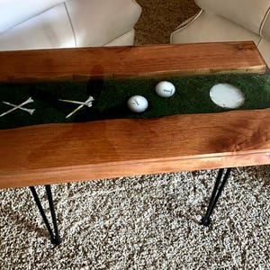 May include: A rectangular coffee table featuring two natural wood slabs with a central river-style inlay of green artificial turf. The inlay contains two white golf balls, four white golf tees, and a circular white golf hole cup, supported by black metal legs.
