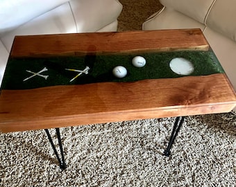 Puede incluir: Una mesa de centro rectangular con dos tablones de madera natural y una incrustación central de césped artificial verde. La incrustación contiene dos pelotas de golf blancas, cuatro tees de golf blancos y un hoyo de golf circular blanco sobre patas de metal.