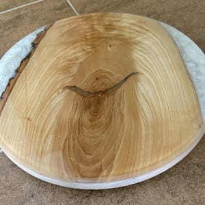 May include: A circular serving board featuring a natural light-colored wood slab with a prominent grain pattern and a rough bark edge on one side, encased in a smooth, translucent white resin border that creates a seamless, polished round finish.