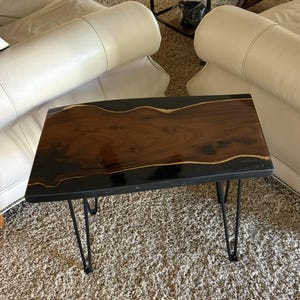 May include: A rectangular side table featuring a live-edge wood slab center with a dark brown grain, encased in a smooth, glossy black resin border. The table is supported by four thin, black metal hairpin legs.
