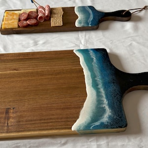 May include: A wooden serving board with a blue and white epoxy resin wave design. The board has a black handle and a leather strap.