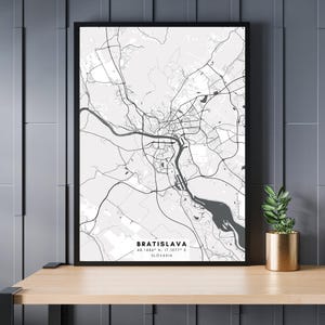 May include: A black and white map print of Bratislava, Slovakia. The map shows the city's streets and waterways. The city's name and coordinates are printed at the bottom of the map.