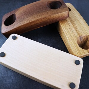 May include: Three wooden coasters with rounded edges and a single hole in the center. The coasters are made of different types of wood: walnut, oak, and maple. Each coaster has four black rubber feet on the bottom.