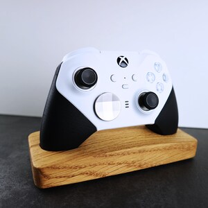 May include: A white Xbox controller with black accents is displayed on a wooden stand. The controller has a white faceplate with a black Xbox logo and a black D-pad.