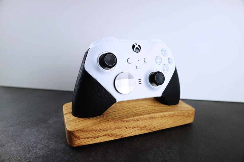 May include: A white Xbox controller with black accents is displayed on a wooden stand. The controller has a white faceplate with a black Xbox logo and a black D-pad.