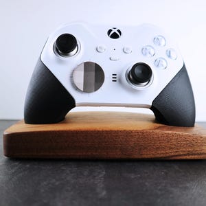 May include: A white Xbox controller with black grips and a custom faceplate is displayed on a wooden stand.