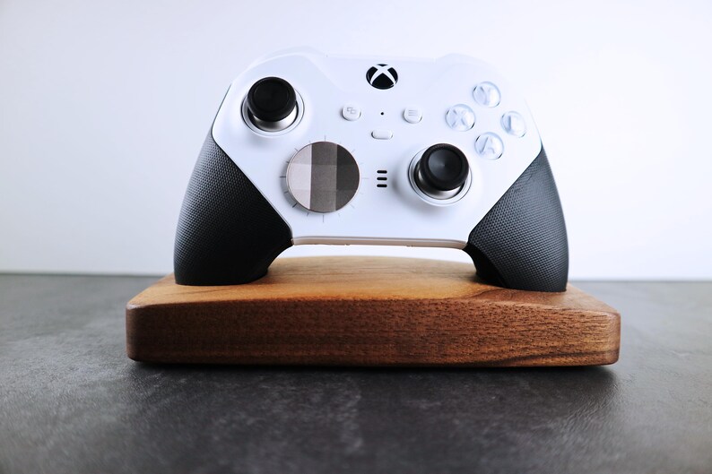 May include: A white Xbox controller with black grips and a custom faceplate is displayed on a wooden stand.
