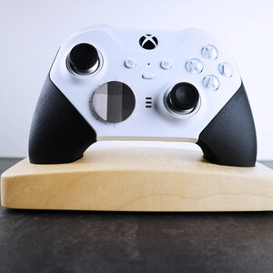 May include: A white Xbox controller with black accents is displayed on a wooden stand. The controller has a textured grip and a circular button in the center.