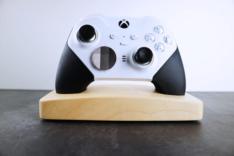 May include: A white Xbox controller with black accents is displayed on a wooden stand. The controller has a textured grip and a circular button in the center.