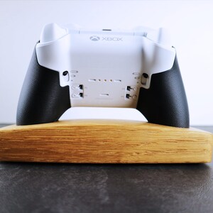 May include: A white Xbox controller with black grips is displayed on a wooden stand. The controller has the word "Xbox" printed on the front.