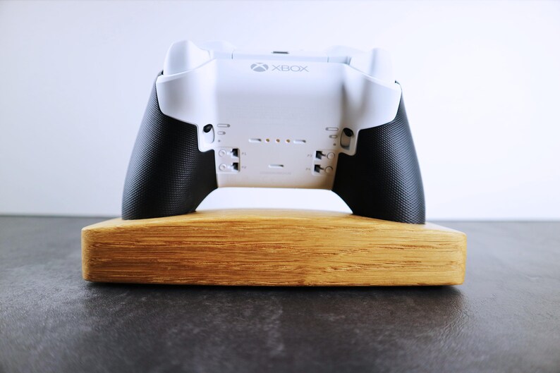 May include: A white Xbox controller with black grips is displayed on a wooden stand. The controller has the word "Xbox" printed on the front.