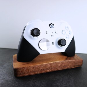 May include: A white Xbox controller with black accents is displayed on a wooden stand. The controller has a white faceplate with a black Xbox logo and black buttons.