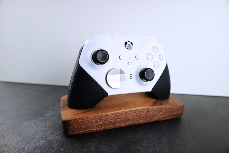May include: A white Xbox controller with black accents is displayed on a wooden stand. The controller has a white faceplate with a black Xbox logo and black buttons.
