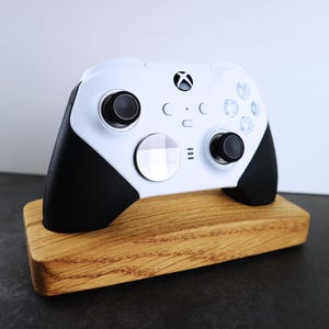 Könnte beinhalten: Ein weißer Xbox-Controller mit schwarzen Akzenten wird auf einem Holzständer präsentiert. Der Controller hat eine weiße Frontplatte mit einem silbernen Knopf und einem schwarzen Knopf auf der rechten Seite.