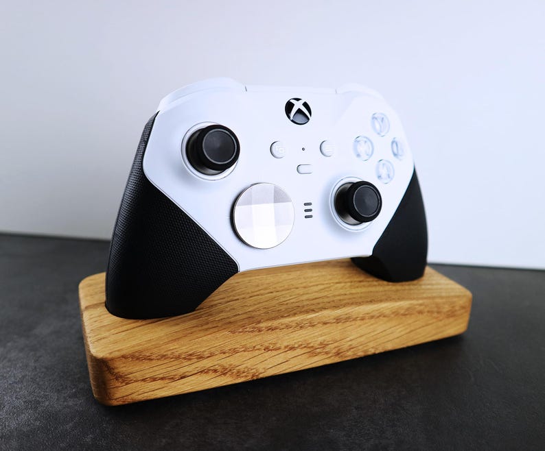May include: A white Xbox controller with black accents is displayed on a wooden stand. The controller has a white faceplate with a silver button and a black button on the right side.