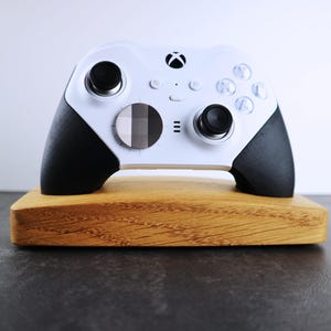 May include: A white Xbox controller with black accents is displayed on a wooden stand. The controller has a black and white patterned button on the front.