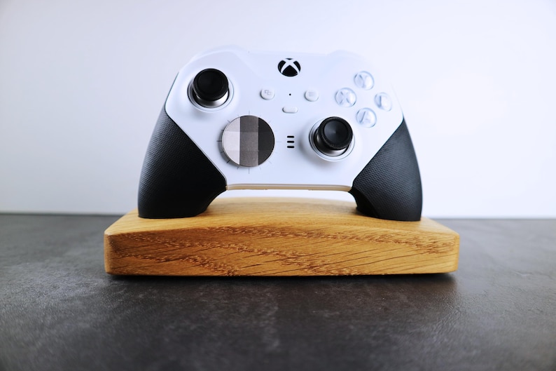 May include: A white Xbox controller with black accents is displayed on a wooden stand. The controller has a black and white patterned button on the front.
