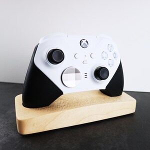 May include: A white Xbox controller with black accents is displayed on a wooden stand. The controller has a white faceplate with a checkered pattern.