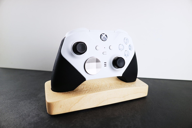 May include: A white Xbox controller with black accents is displayed on a wooden stand. The controller has a white faceplate with a checkered pattern.