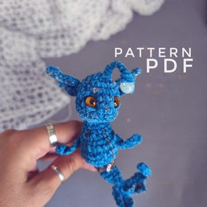 Könnte beinhalten: Eine handgefertigte blaue Häkelpuppe mit großen Augen und einem Schwanz. Die Puppe hat kleine Arme und Ohren sowie eine kleine, klare Perle auf dem Kopf. Der Text "PATTERN PDF" ist in Weiß sichtbar. Die Puppe wird von einer Person gehalten.
