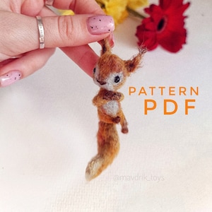 Peut inclure: Gros plan sur un petit jouet en tricot de forme d'écureuil brun et blanc tenu par une main. Le texte "PATTERN PDF" est écrit en lettres orange sur fond blanc. Le texte "@mavdrik_toys" est écrit en lettres noires sur fond blanc.