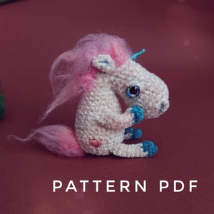 May include: A crocheted unicorn amigurumi toy with pink yarn hair, blue eyes, and a blue horn. The unicorn is sitting on a red surface with the text "PATTERN PDF" below it.